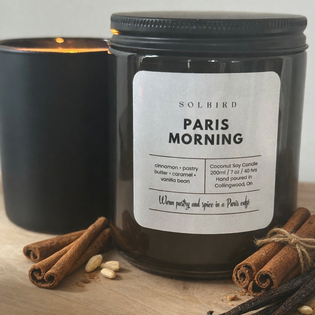 Black candle jar labeled 'Paris Morning' with cinnamon sticks and vanilla beans on a wooden surface.